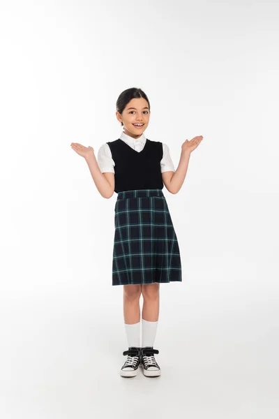 Full length, cheerful schoolkid in uniform gesturing and looking at camera on white, girl in skirt — Stock Photo