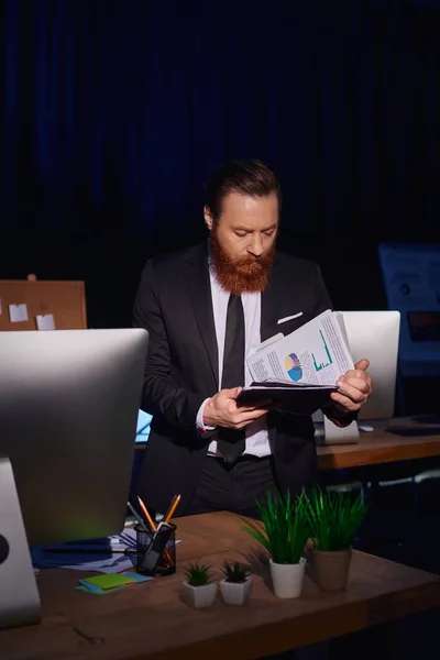Бородатый бизнесмен в костюме работает допоздна и просматривает документы с инфографикой в ночном офисе — стоковое фото