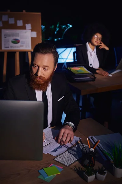 Бородатый мужчина, работающий с документами и компьютером рядом с очаровательной африканской женщиной на заднем плане — стоковое фото