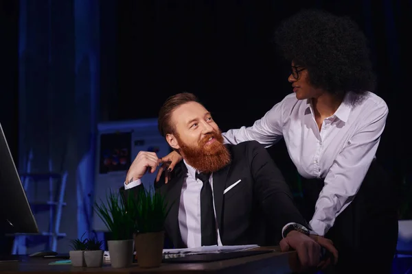 Афроамериканська жінка обіймає щасливого бородатого бізнесмена в нічному офісі, любовний роман на роботі — стокове фото