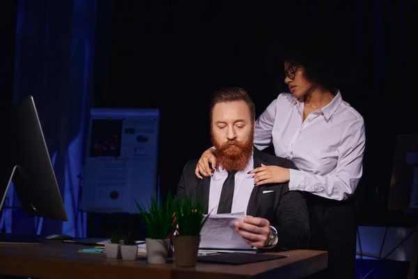 Séduisante femme afro-américaine embrassant les épaules d'un homme d'affaires sérieux travaillant avec des papiers — Photo de stock