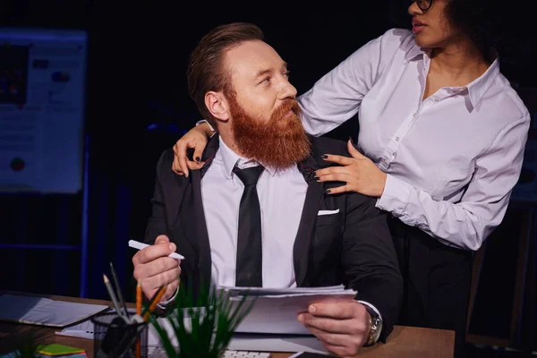Homme d'affaires barbu positif avec des papiers près de charmante femme afro-américaine flirter dans le bureau — Photo de stock