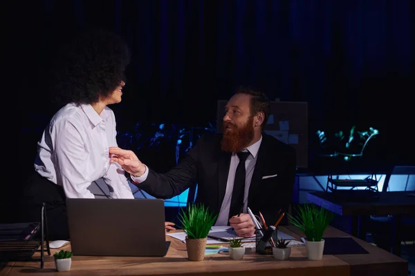 Бородатый бизнесмен трогает блузку соблазнительной африканской американки возле ноутбука в ночном офисе — стоковое фото