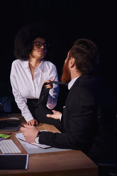 Mujer afroamericana en gafas tirando de corbata de hombre de negocios barbudo en la noche, romance de oficina - foto de stock