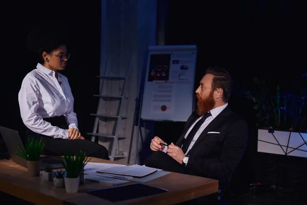 Bearded businessman looking at pretty african american woman sitting on work desk at night in office — Stock Photo