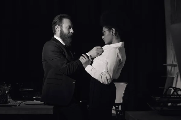 Black and white photo of businessman undressing african american woman in night office, side view — Stock Photo
