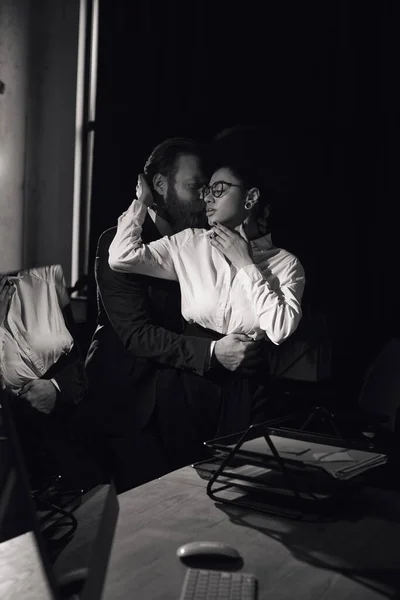 Couple interracial en tenue formelle embrassant dans le bureau la nuit, histoire d'amour, photo noir et blanc — Photo de stock