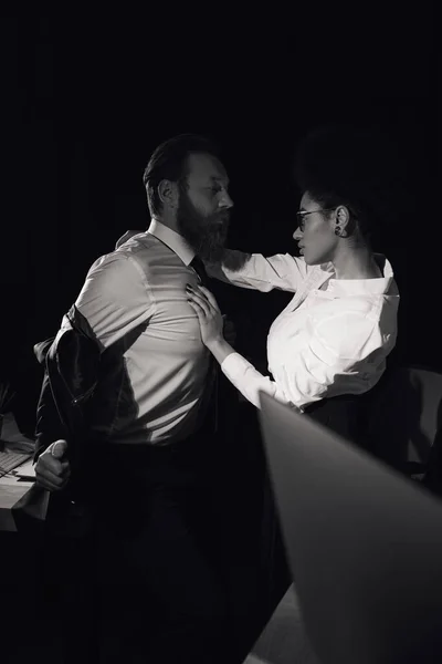 Femme afro-américaine passionnée embrassant homme d'affaires la nuit au bureau, photo noir et blanc — Photo de stock