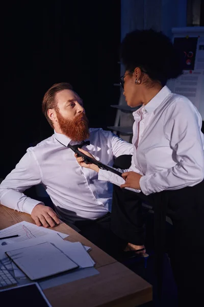 Mujer afroamericana tirando de corbata de hombre de negocios barbudo en la oficina nocturna, amorío en el trabajo - foto de stock