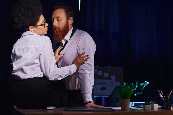 Mulher americana africana atraente seduzindo homem de negócios barbudo na mesa de trabalho no escritório da noite, romance — Fotografia de Stock