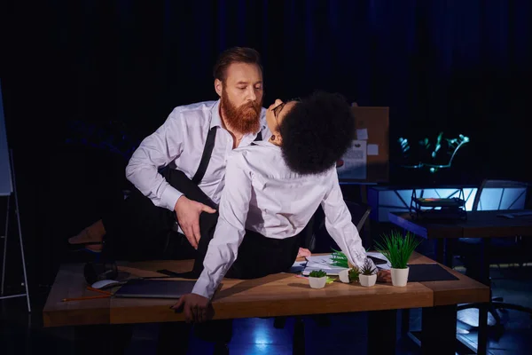 Bearded businessman embracing young african american woman on work desk in dark office, secret love — Stock Photo