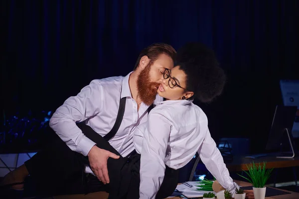 Homme d'affaires barbu étreignant jeune femme afro-américaine dans des lunettes pendant le quart de nuit au bureau — Photo de stock