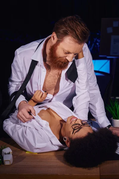 Hombre de negocios barbudo desnudando mujer afroamericana feliz en el escritorio de trabajo en la oficina nocturna, seducción - foto de stock