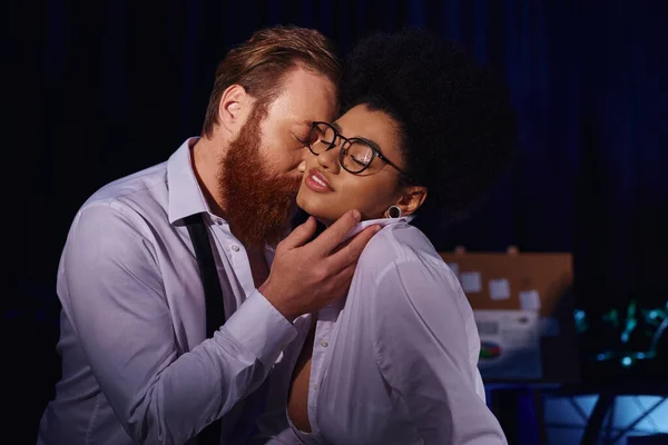 Bearded businessman kissing smiling african american woman in eyeglasses, office romance at night — Stock Photo