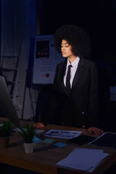 Jovem mulher de negócios afro-americana olhando para monitor de computador perto de documentos no escritório noturno — Fotografia de Stock