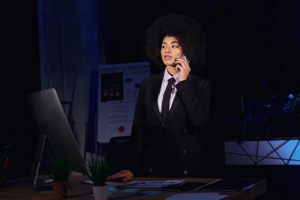 African american businesswoman talking on smartphone near computer monitor in office at night — Stock Photo