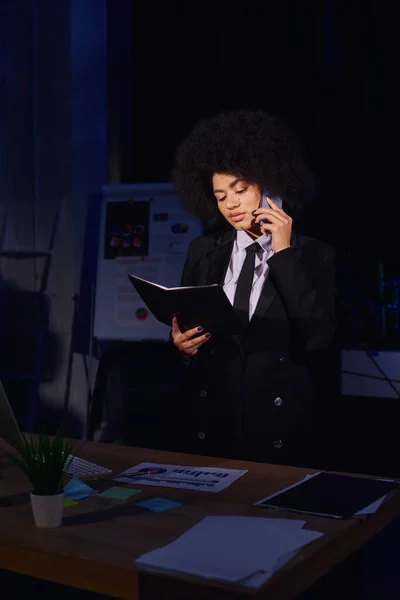 Mulher de negócios afro-americana com notebook falando no smartphone enquanto trabalhava no escritório à noite — Fotografia de Stock