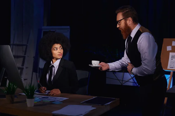 Bärtiger Geschäftsmann hält Kaffee in der Nähe einer afrikanisch-amerikanischen Sekretärin und flirtet im Nachtbüro — Stockfoto