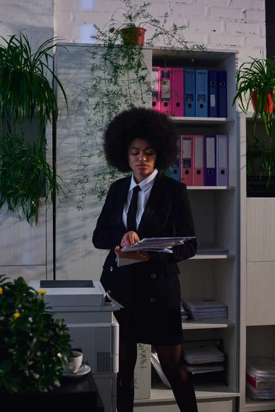 Mulher afro-americana em desgaste formal de pé com documentos em papel perto copiadora à noite no escritório — Fotografia de Stock