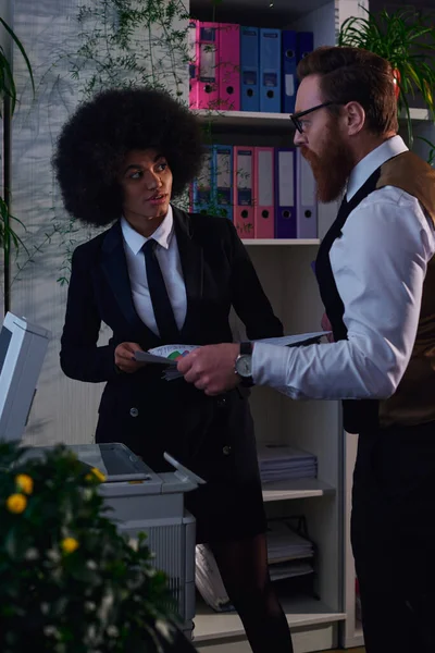Hombre de negocios barbudo con documentos que explican a la mujer afroamericana cómo usar una fotocopiadora en la oficina - foto de stock