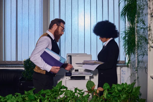 Hombre de negocios barbudo con colega afroamericano copiando documentos en la fotocopiadora en la oficina, vista lateral - foto de stock