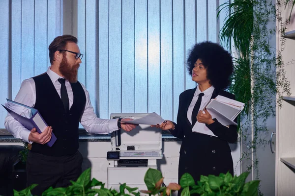 Homem de negócios barbudo em óculos dando documentos para jovem secretário afro-americano perto copiadora — Fotografia de Stock