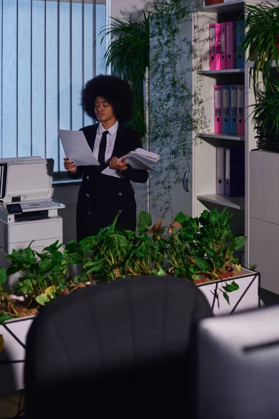 Femme d'affaires afro-américaine avec des documents près du copieur et des plantes décoratives la nuit au bureau — Photo de stock