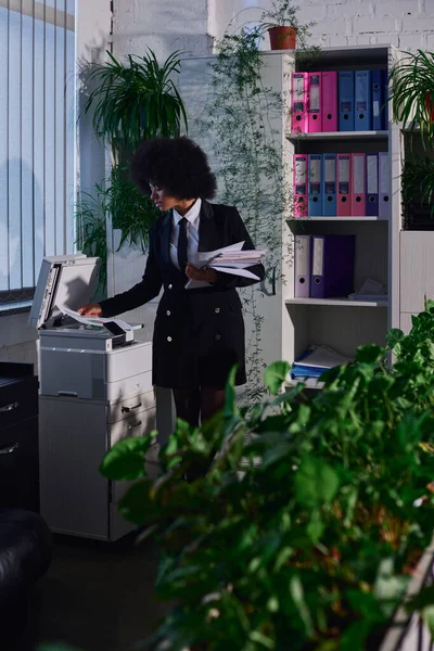 Jovem mulher afro-americana copiando documentos na máquina copiadora à noite no escritório, excesso de trabalho — Fotografia de Stock