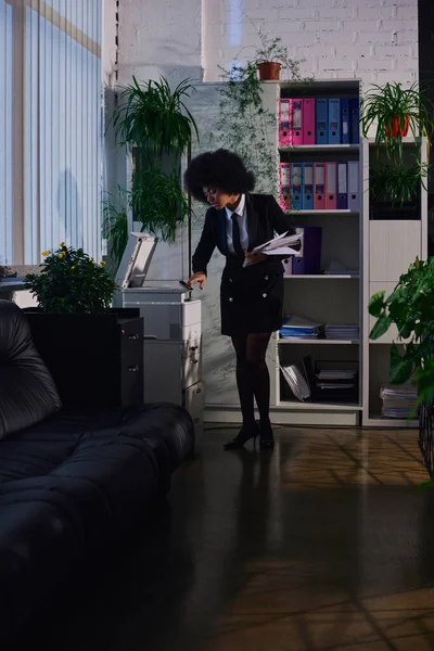 Mujer afroamericana en ropa formal elegante copiar documentos mientras trabaja en la oficina por la noche - foto de stock