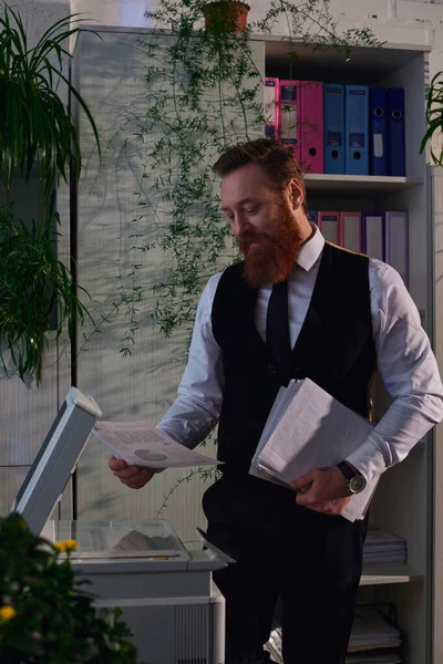 Hombre de negocios barbudo de pie con documentos cerca de la fotocopiadora mientras trabaja en la oficina por la noche - foto de stock