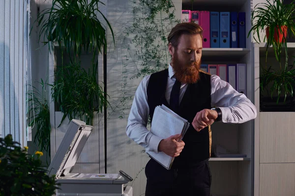 Homme d'affaires barbu avec des papiers regardant montre-bracelet près copieur tout en travaillant tard dans le bureau — Photo de stock