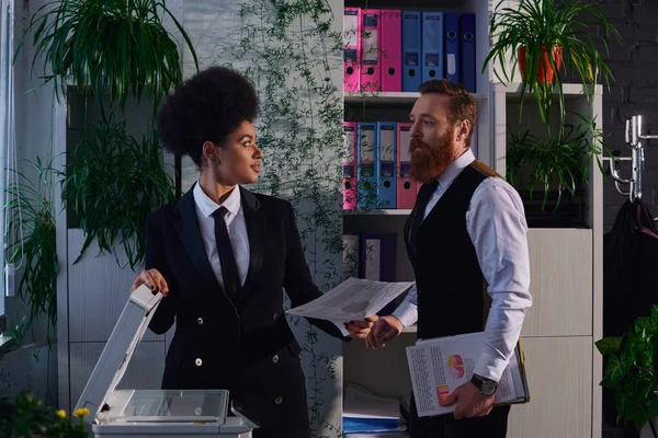 Colegas interraciales con documentos mirándose unos a otros cerca de la fotocopiadora en la oficina por la noche - foto de stock