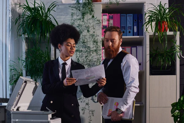 Joven secretaria afroamericana mostrando documento a empresario barbudo cerca de fotocopiadora en oficina - foto de stock