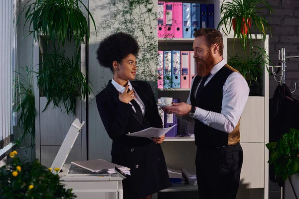 Homme d'affaires barbu tenant tasse de café et flirtant avec le secrétaire afro-américain au bureau — Photo de stock