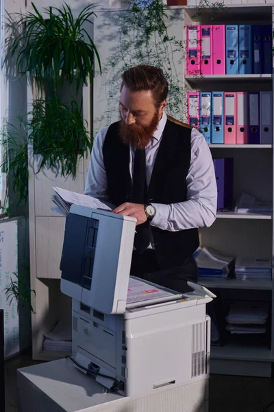 Hombre de negocios barbudo en ropa formal mirando documentos cerca de la fotocopiadora mientras trabaja hasta tarde en la oficina - foto de stock