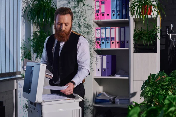 Hombre de negocios barbudo enfocado fotocopiando documentos en fotocopiadora en la noche en la oficina, sobrecarga de trabajo - foto de stock