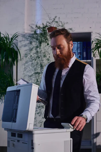 Homem de negócios barbudo focado copiar documentos na máquina copiadora enquanto trabalhava até tarde no escritório — Fotografia de Stock