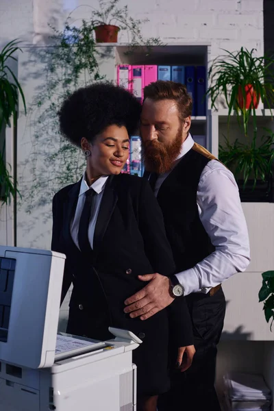 Uomo d'affari barbuto abbracciando sorridente donna africana americana vicino copiatrice, seduzione in ufficio — Foto stock