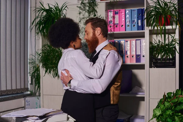 Casal multiétnico em desgaste formal abraçando no escritório perto de documentos em papel à noite, sedução — Fotografia de Stock