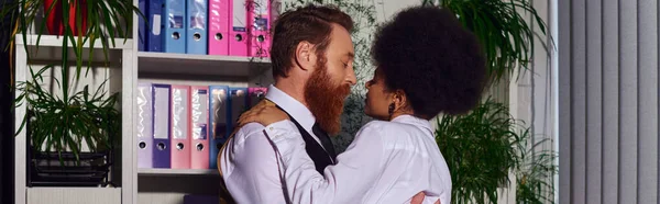 Interracial couple embracing  in evening near rack with folders in evening, love at work, banner — Stock Photo