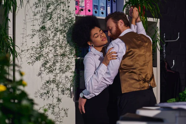 Passionate interracial couple embracing near decorative plants in office at night, work romance — Stock Photo