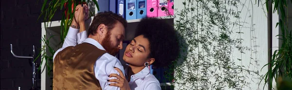 Passionate interracial couple embracing near decorative plants in office at night, banner — Stock Photo