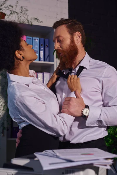 Young african american secretary seducing and undressing bearded businessman in office at night — Stock Photo
