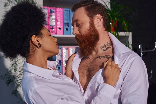 Apasionada mujer afroamericana desnudándose barbudo hombre de negocios tatuado, romance en la oficina nocturna - foto de stock