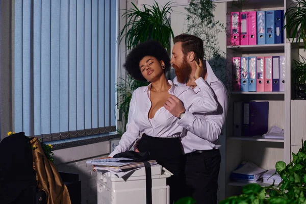 Empresário barbudo seduzindo mulher americana africana quente perto copiadora e documentos no escritório à noite — Fotografia de Stock