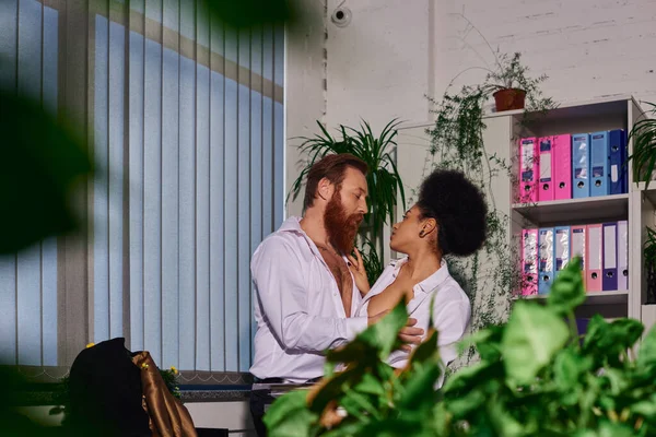 Passionate interracial couple embracing in night office on blurred foreground, love affair at work — Stock Photo