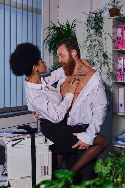 Amor y afecto, hombre de negocios tatuado abrazando a la joven afroamericana en la oficina nocturna - foto de stock