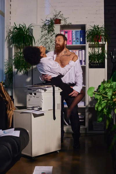 Passion et séduction, homme d'affaires barbu tatoué embrassant femme chaude afro-américaine près de copieur — Photo de stock