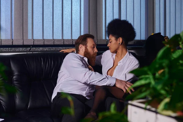 Sexy afroamericana mujer de negocios coqueteando con barbudo colega en sofá en la oficina por la noche - foto de stock