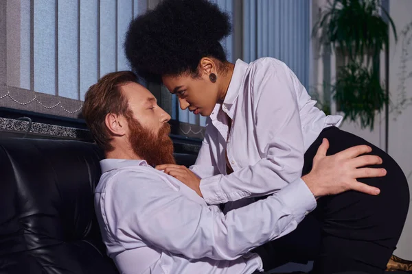 Соблазнительная африканская американская секретарша соблазняет бородатого бизнесмена на диване в ночном офисе, вид сбоку — стоковое фото